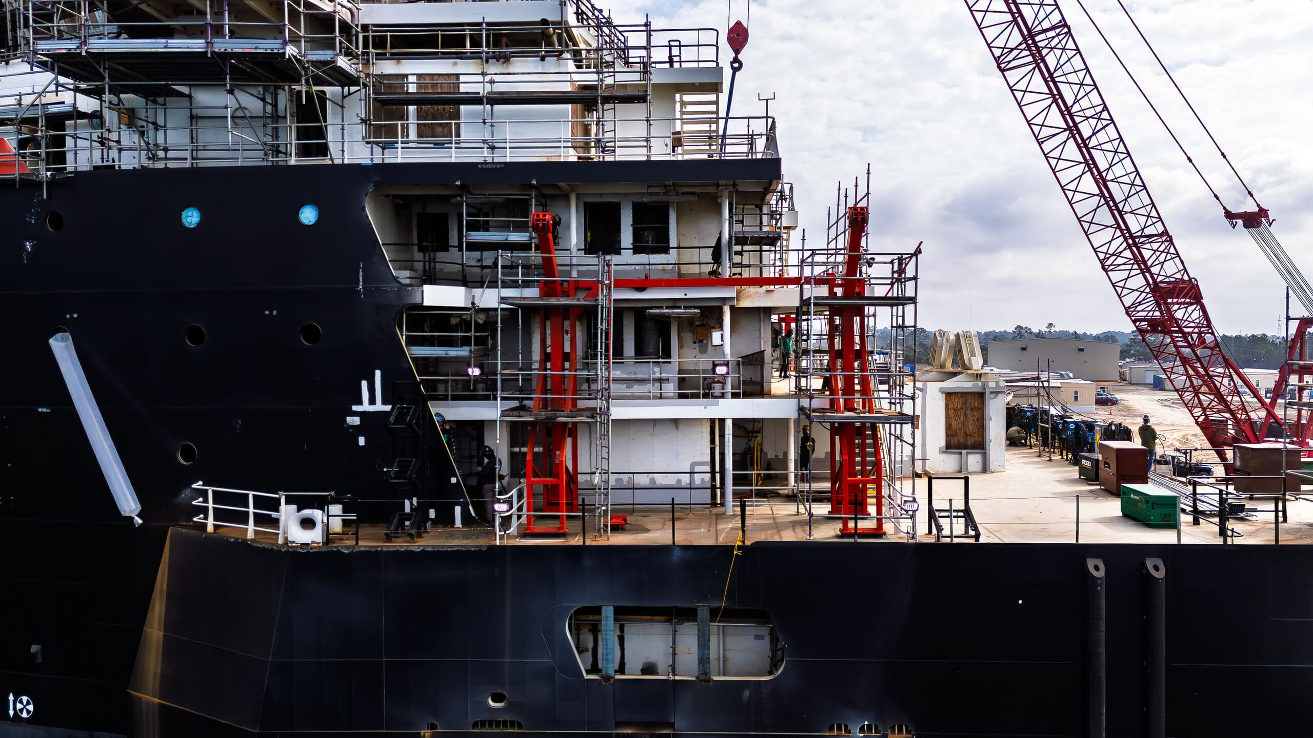 A vessel under construction in the yard, superstructure wrapped in scaffolding beside a crawler crane